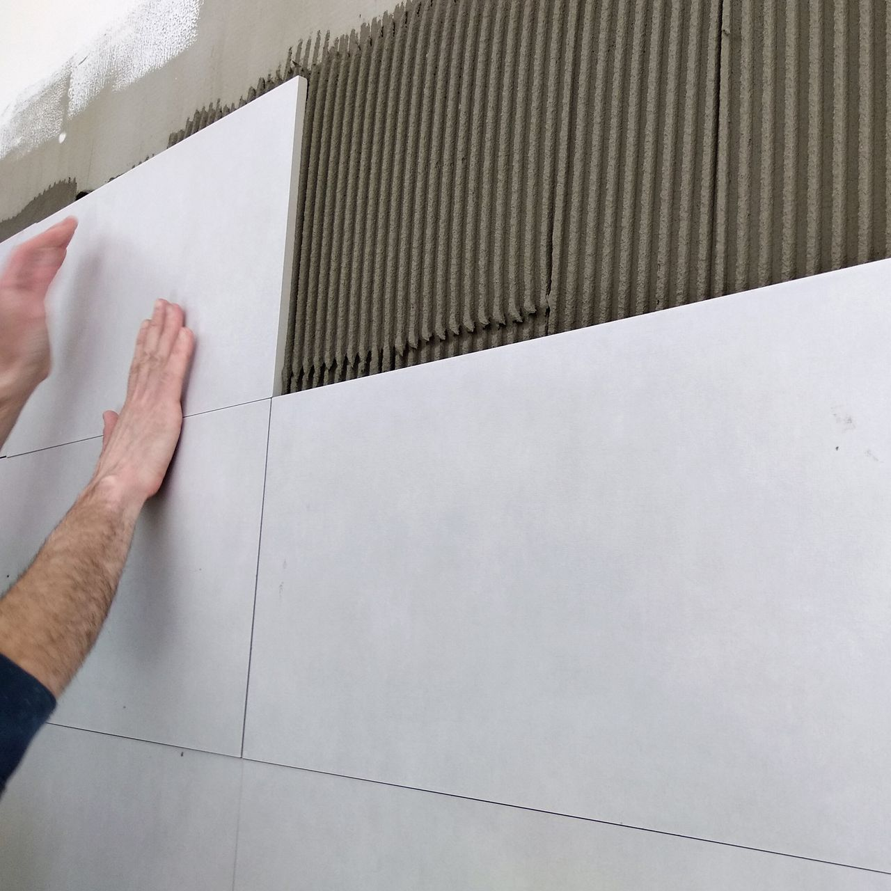 Positionnement manuel des carreaux muraux blancs sur l'adhésif gris.