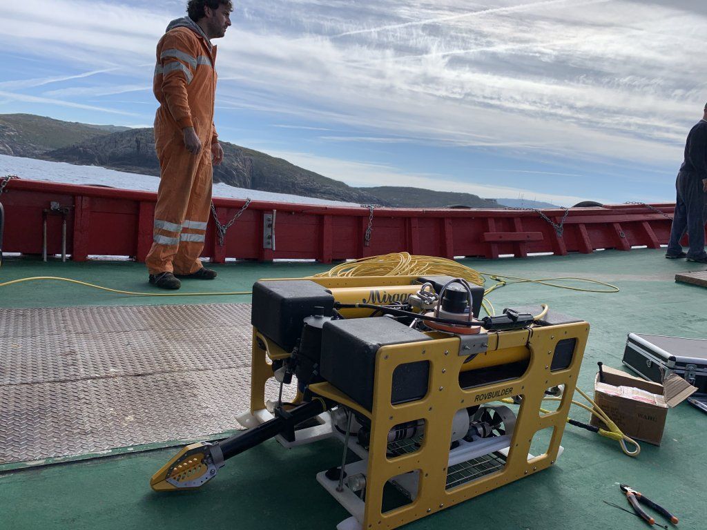 Dron submarino amarillo en cubierta con una persona en un mono naranja; fondo de océano y cielo.