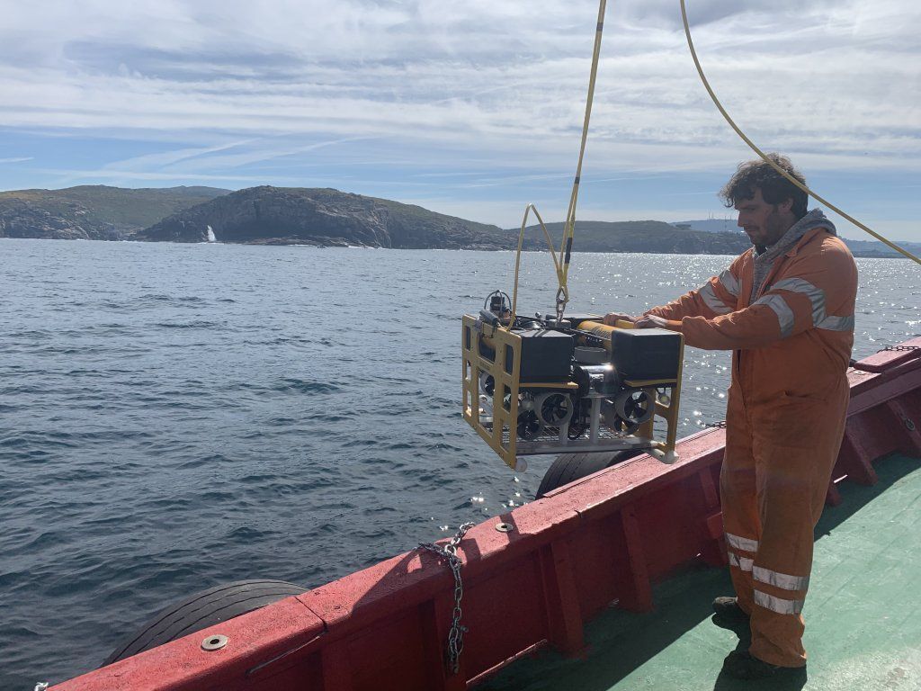 Un hombre con un mono naranja en un barco baja un dron submarino con forma de jaula amarilla hacia el mar.