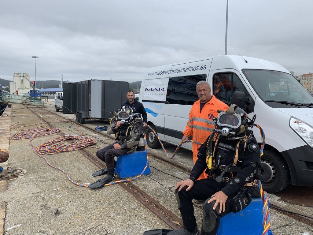 Dos buzos equipados se sientan en barriles azules cerca de una camioneta con una tercera persona, muelles y equipo.