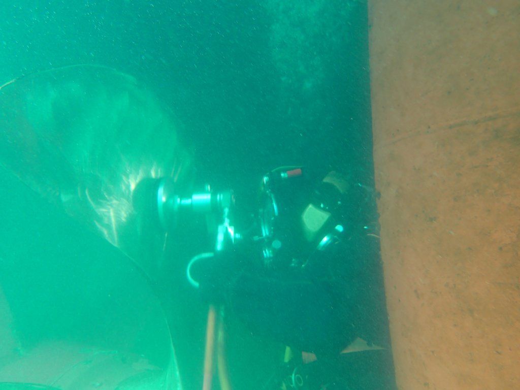 Buceador bajo el agua, con una cámara en la mano, examina una estructura. Una luz verde ilumina la escena, aguas oscuras.