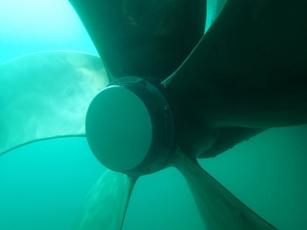 Vista submarina de una hélice de barco con tres palas, el agua es de color azul verdoso.