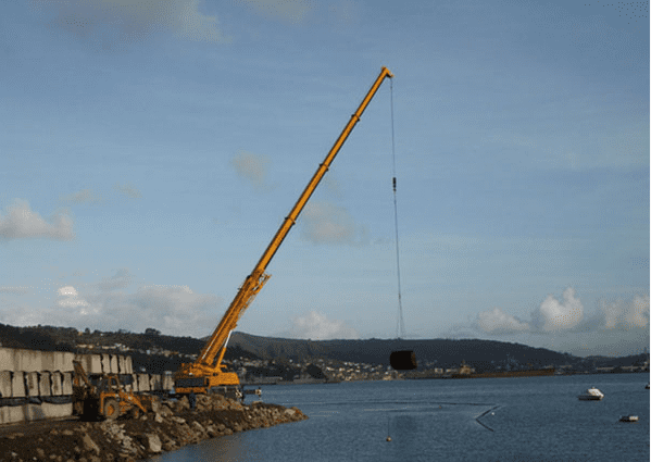 Una grúa amarilla levanta un objeto oscuro sobre el agua, junto a una estructura de hormigón. Cielo azul y agua.