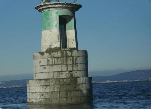 Faro en el mar: estructura de torre verde y blanca con una puerta, contra un cielo azul y agua.