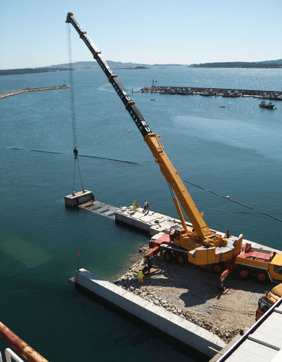 Grúa elevando una estructura de hormigón en un puerto. Trabajadores en la estructura y a lo largo de la orilla. Agua azul.
