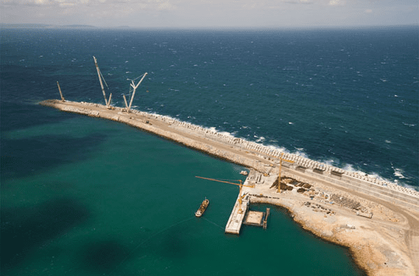 Construcción de un muelle costero con grúas, agua turquesa y olas rompientes.