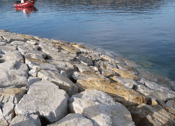 Costa rocosa con piedras de colores claros, agua azul tranquila y un barco rojo en la distancia.