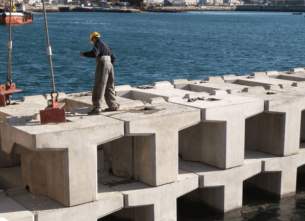 Obrero de la construcción con casco amarillo sobre un rompeolas de hormigón, cerca del agua, con una grúa encima.