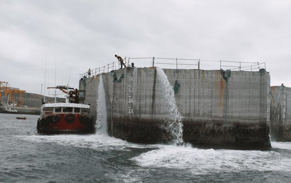 Agua brotando de una estructura de hormigón hacia el mar; cerca hay un pequeño bote con una manguera. Cielo nublado.