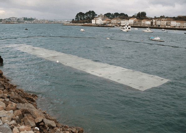 Una plataforma rectangular de hormigón sumergida en el agua cerca de una orilla rocosa, con barcos y edificios al fondo.