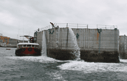 Un gran cajón de hormigón se inunda junto a un barco en el océano. El agua se desborda por los laterales.