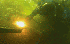 Buzo soldando bajo el agua con chispas de color amarillo brillante, vistiendo equipo de buceo en aguas turbias.