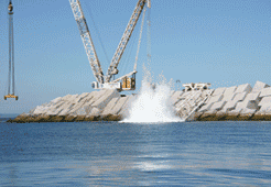 Grúa arrojando un bloque de hormigón al océano, creando un gran chapoteo cerca de un rompeolas.