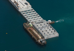 Barcazas en el agua, una grande y en forma de cuadrícula, junto a otra más pequeña. Un pequeño bote se mueve cerca.