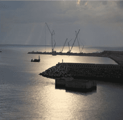 Grúas trabajando en un rompeolas en un puerto. La luz del atardecer se refleja en las tranquilas aguas.