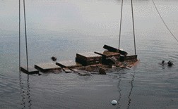 Una plataforma rectangular sumergida con equipaje encima se levanta del agua mediante cuerdas.