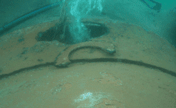 Vista submarina de un objeto metálico corroído con una abertura circular y un detalle en forma de arco, con burbujas ascendiendo.
