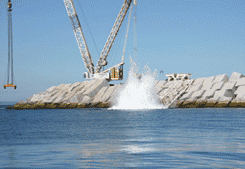 Grúa dejando caer un bloque de hormigón al agua, provocando un gran chapoteo cerca de un rompeolas.