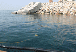Agua azul, un muelle hecho de bloques de hormigón apilados y rocas, con una boya amarilla flotando.