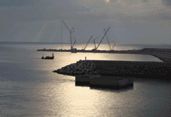 Grúas de construcción trabajando en un rompeolas que se extiende hacia el océano bajo un cielo nublado.