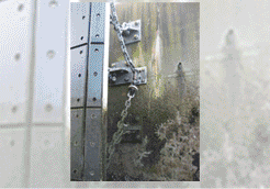 Puerta metálica con cadena y pestillo. La puerta está fijada a una pared musgosa y erosionada.