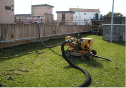 Bomba amarilla con mangueras sobre el césped, cerca de un muro de hormigón y edificios.