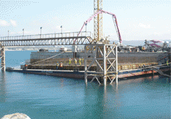 Obreros de la construcción vierten hormigón en una barcaza en el mar con una grúa y una pasarela.