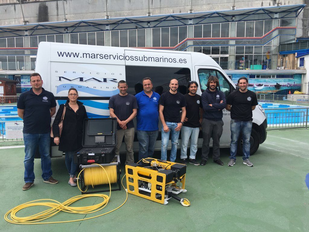 Grupo de personas posando con una furgoneta y equipo subacuático al aire libre cerca de una piscina.