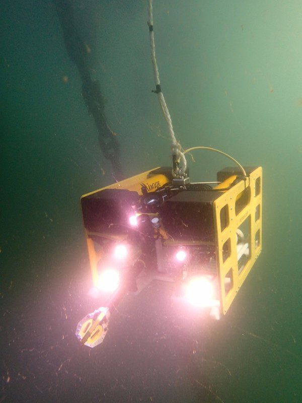 ROV submarino amarillo con luces brillantes, suspendido de un cable en aguas turbias.
