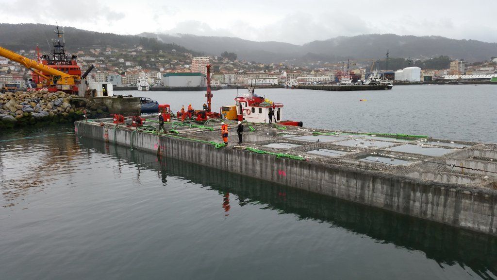 Barcaza de construcción en un puerto con maquinaria y trabajadores; ciudad y montañas al fondo.