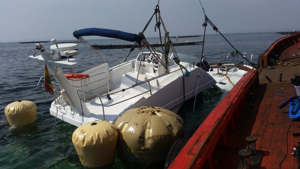 Un velero blanco se saca del agua con una grúa. Cerca hay boyas amarillas y una barcaza roja.