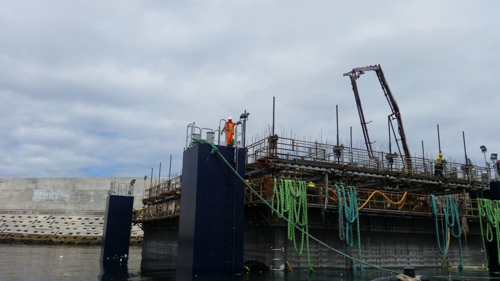 Obra sobre el agua. Trabajadores con trajes naranjas en una estructura azul oscuro, andamios y grúa. Cielo nublado.