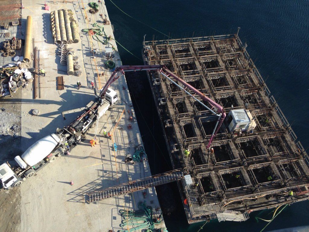 Hormigón vertido en una gran estructura flotante en forma de rejilla en un muelle. Un camión hormigonera bombea cemento.