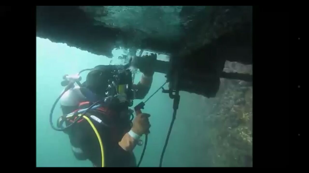 Buzo trabajando bajo el agua, sosteniendo una herramienta, cerca de los restos de un barco en aguas claras.
