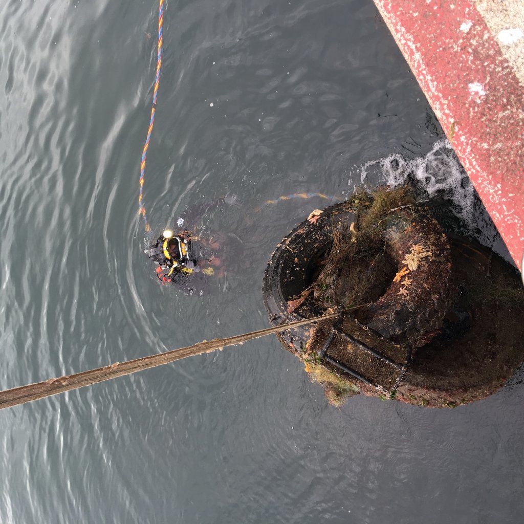 Buceador en el agua cerca de un objeto grande y oxidado y un barco, probablemente durante una inspección o trabajo submarino.