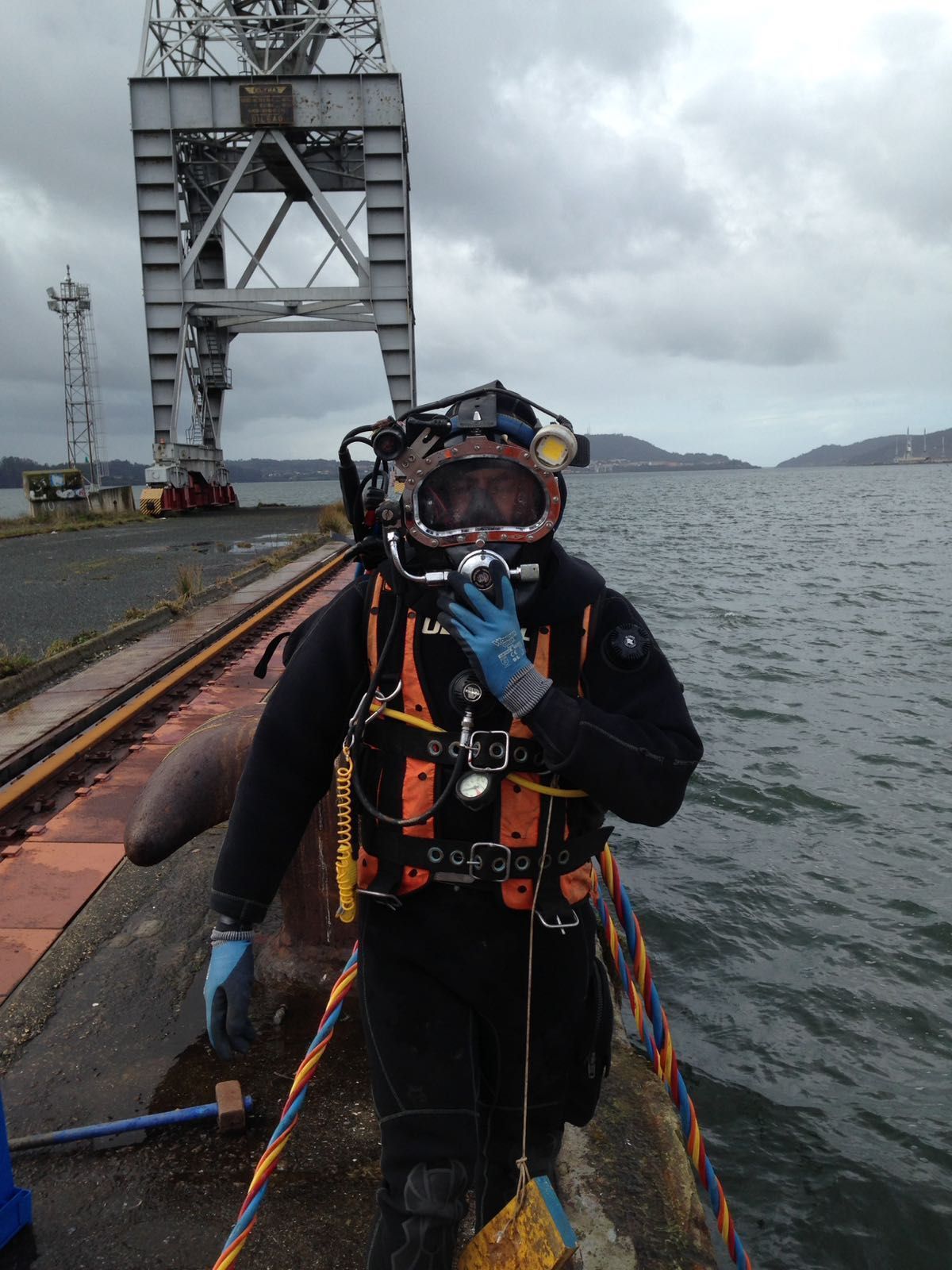 Buzo con traje negro y casco en un muelle. Grúa y agua al fondo, cielo nublado.