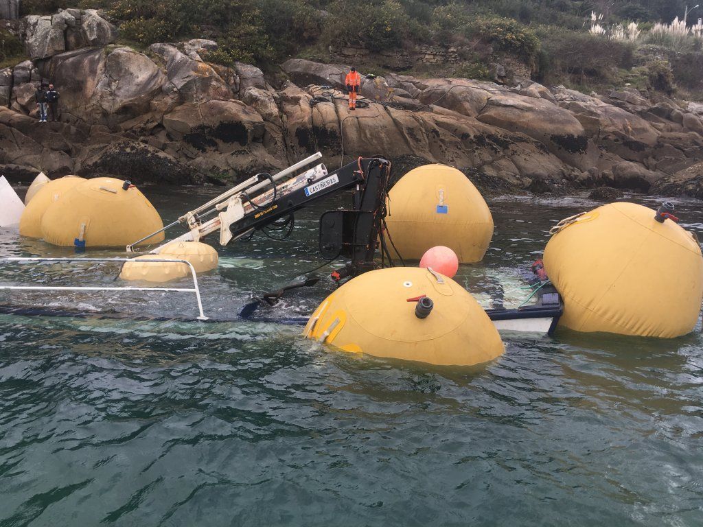 Buque parcialmente sumergido con dispositivos de flotación amarillos cerca de la costa rocosa.
