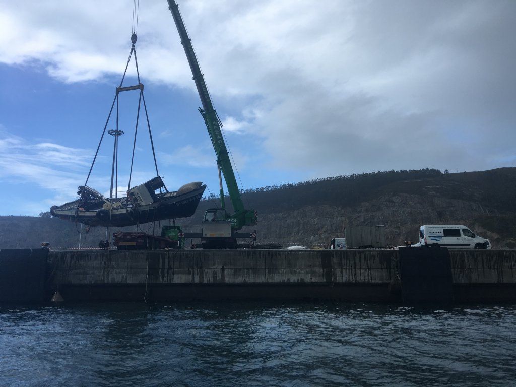 Una grúa levanta un barco averiado fuera del agua en un puerto, con el cielo nublado encima.