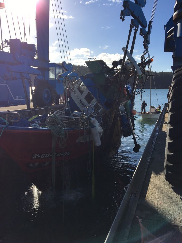 Barco pesquero en el muelle, con el equipo izado. Agua, cielo azul y otros barcos visibles.