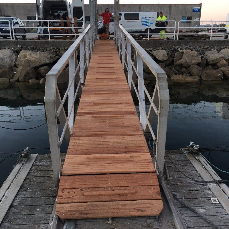 Rampa de muelle de madera con barandilla plateada, que conduce a una persona parada en la parte superior, atracada sobre el agua.