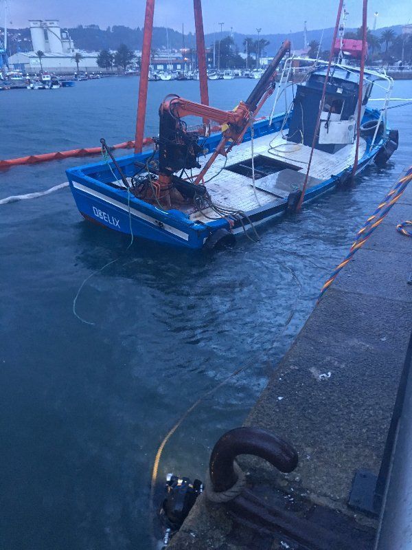 Un barco azul parcialmente sumergido está siendo sacado del agua con una grúa naranja en un puerto.