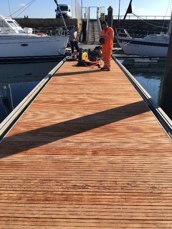 Trabajadores en un muelle de madera cerca de los barcos; uno de ellos con traje naranja utiliza una máquina.