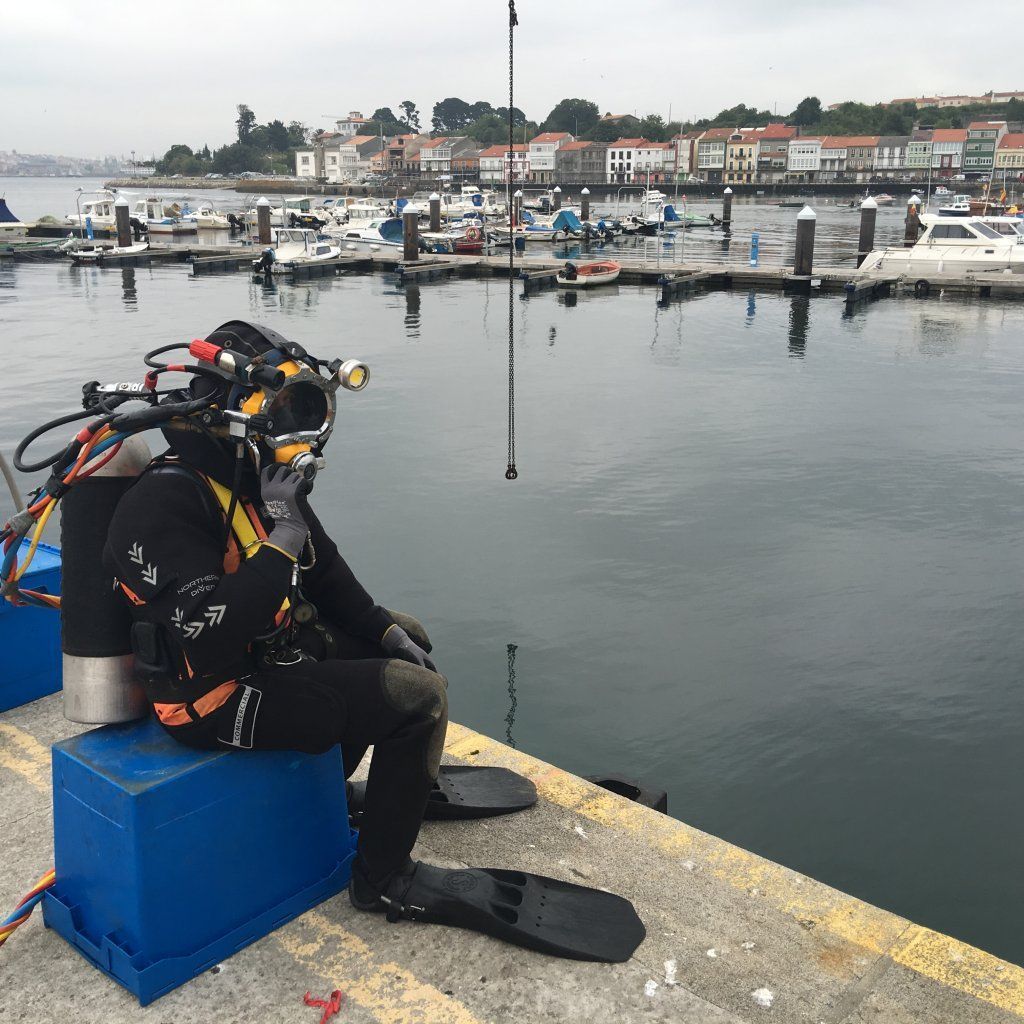 Buzo con equipo completo sentado en una caja azul en el muelle; puerto y edificios en el fondo.