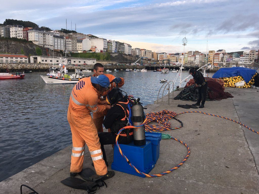 Dos personas con monos de color naranja ayudan a un buceador con traje de neopreno y equipo de buceo en un muelle cerca de un puerto con edificios de la ciudad.