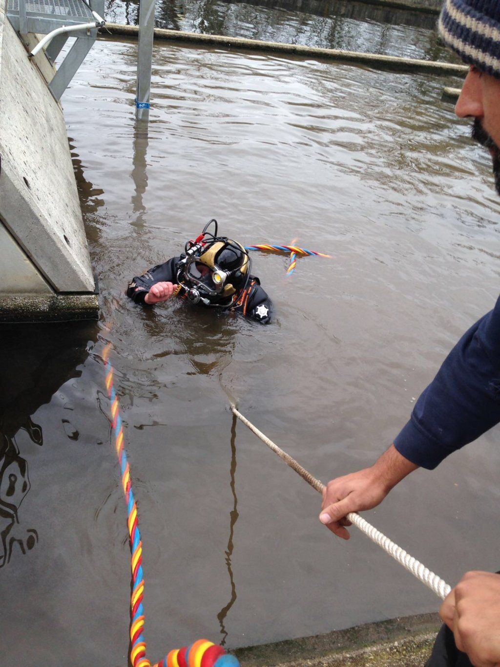 Buzo con equipo negro siendo rescatado del agua por alguien que sujeta una cuerda. Muro de hormigón y soporte al fondo.