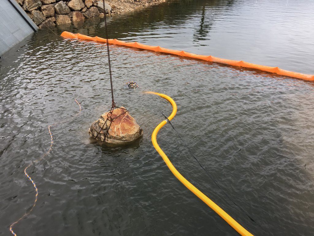 Agua con barrera flotante de color naranja, manguera amarilla y un objeto rocoso parcialmente sumergido.