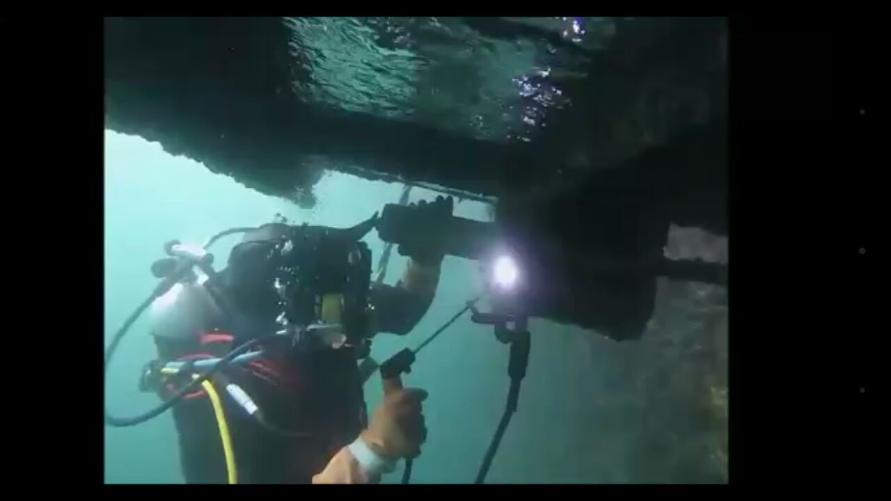 Buzo bajo una estructura, usando una herramienta. La luz ilumina su trabajo. El agua es verdosa.