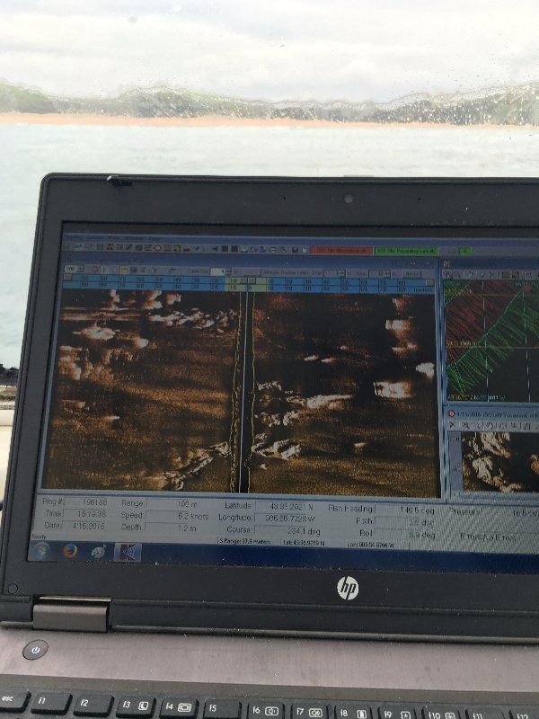 Pantalla de portátil con datos del sonar; barco en el agua. Elementos submarinos marrones y blancos. Fondo de cielo nublado.