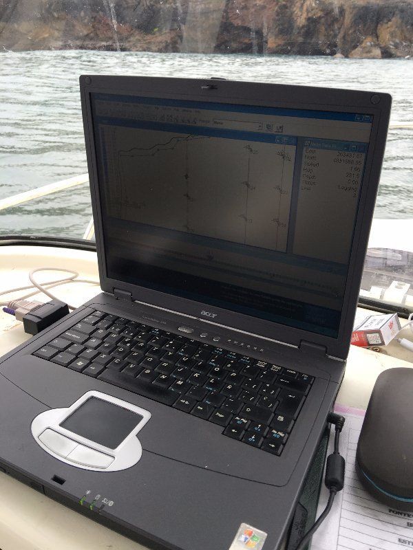 Computadora portátil en la cubierta de un barco mostrando datos; mouse y otros equipos visibles, agua en el fondo.