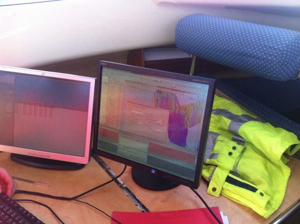 Dos monitores de computadora sobre un escritorio de madera con un chaleco de seguridad amarillo y un cojín azul.